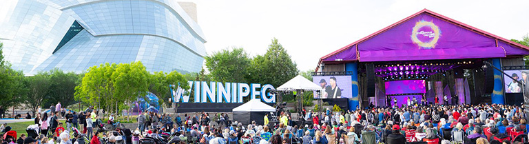 Une foule de gens assis sur des chaises longues regardent un spectacle sur une scne. Un grand panneau de Winnipeg se trouve  l'arrire-plan devant le Muse des droits de la personne.